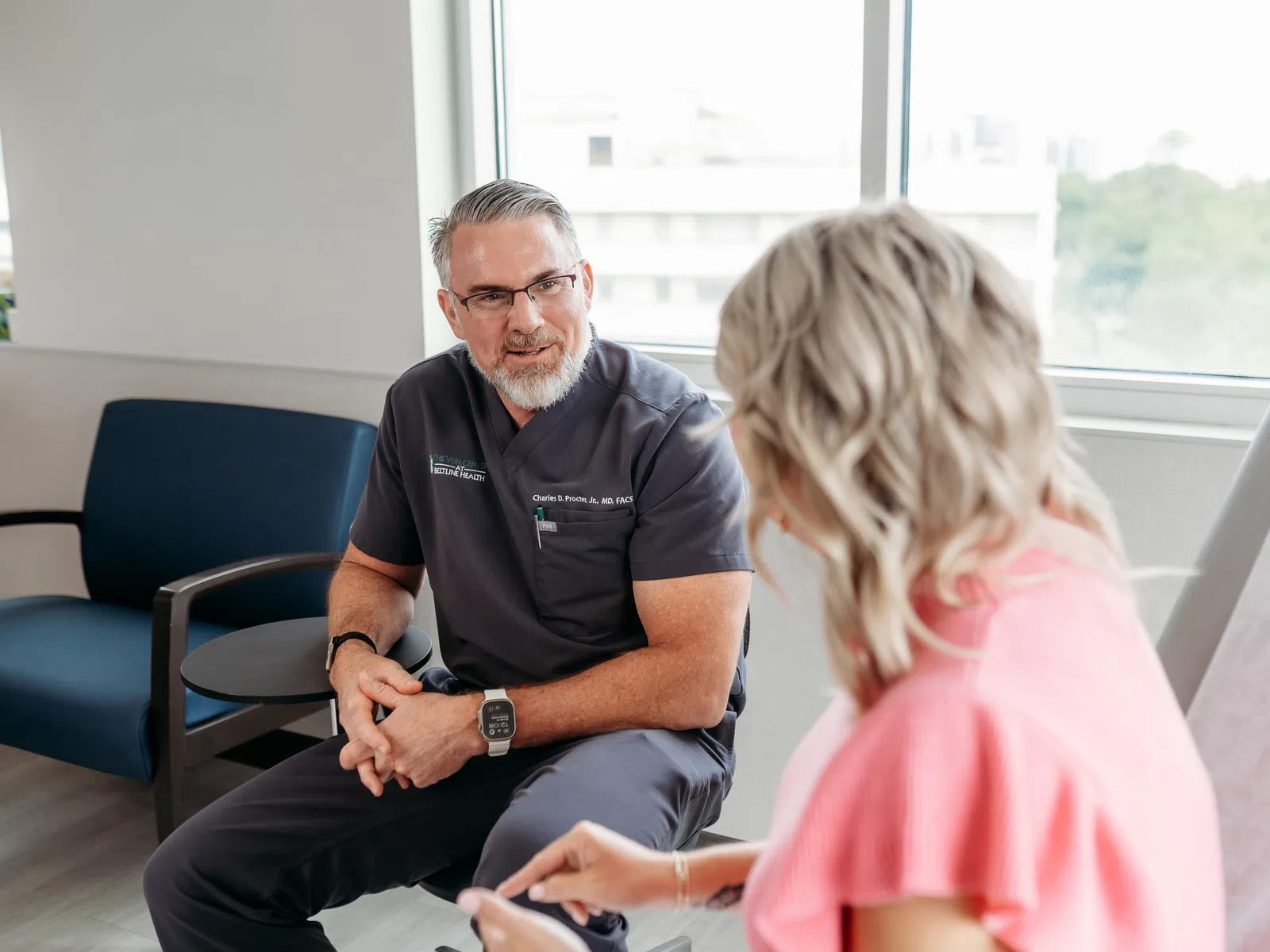 Doctor in scrubs talking to a female patient in a bright medical office with large windows and natural light.