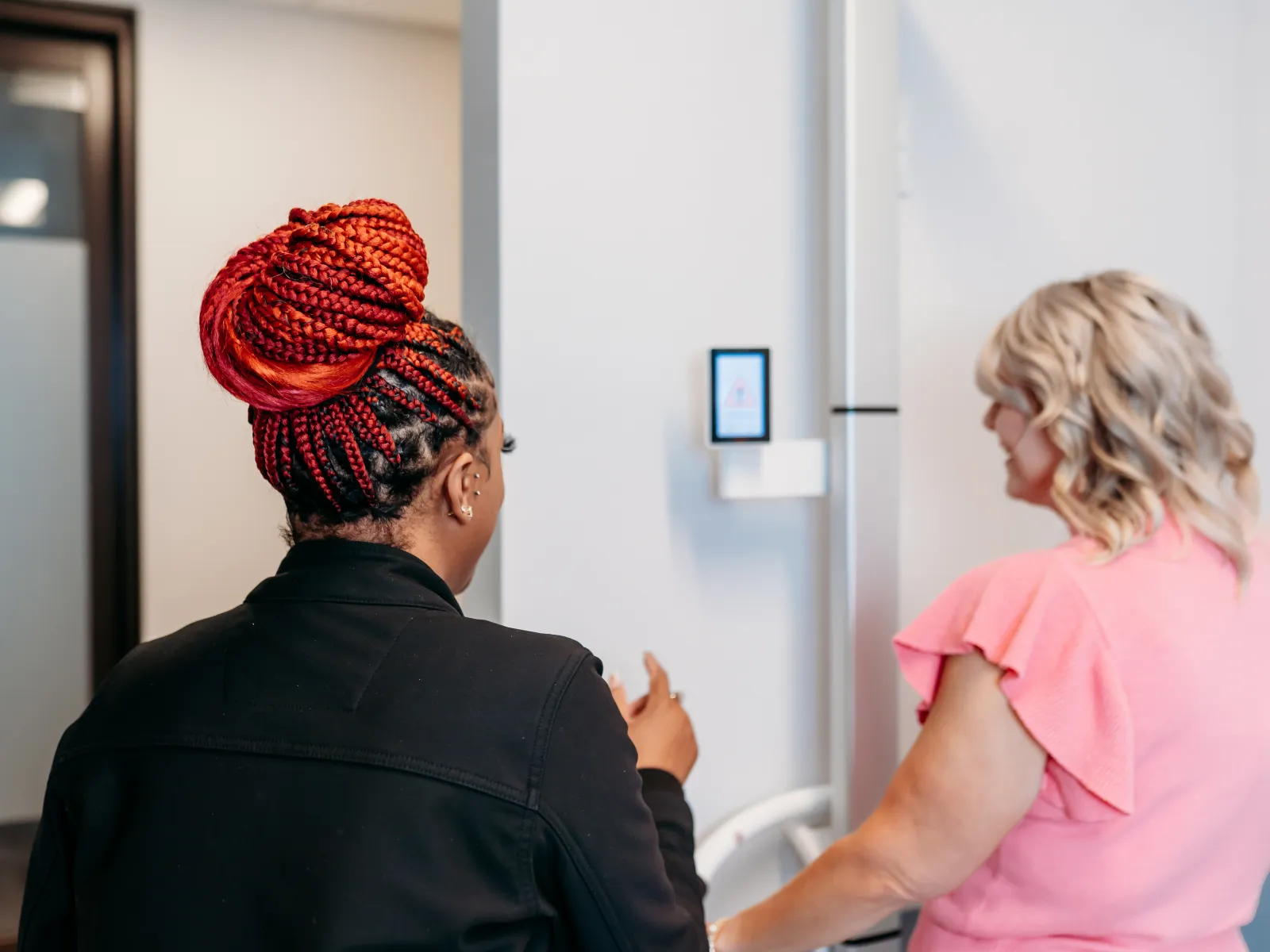 Two women in conversation indoors, one with red braided hair in a bun, the other wearing a pink ruffled sleeve top.