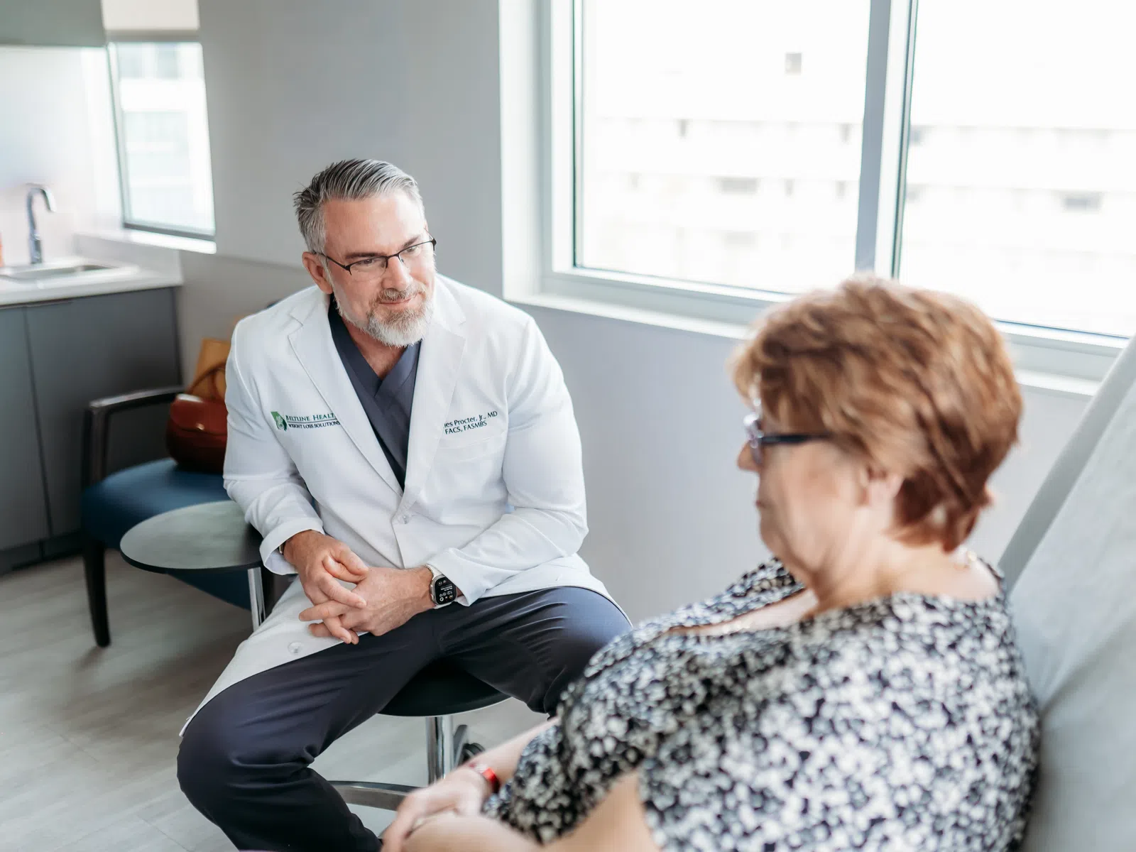 Doctor in white coat consulting with a seated female patient in a modern medical office with natural light.