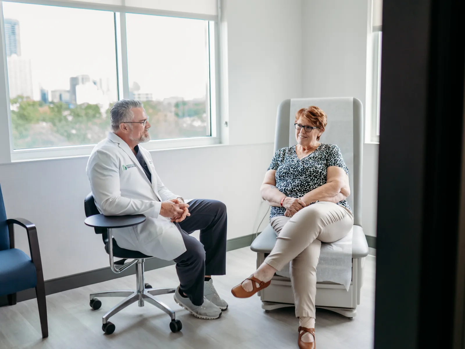 Doctor in white coat consulting with female patient in bright medical office with large windows.