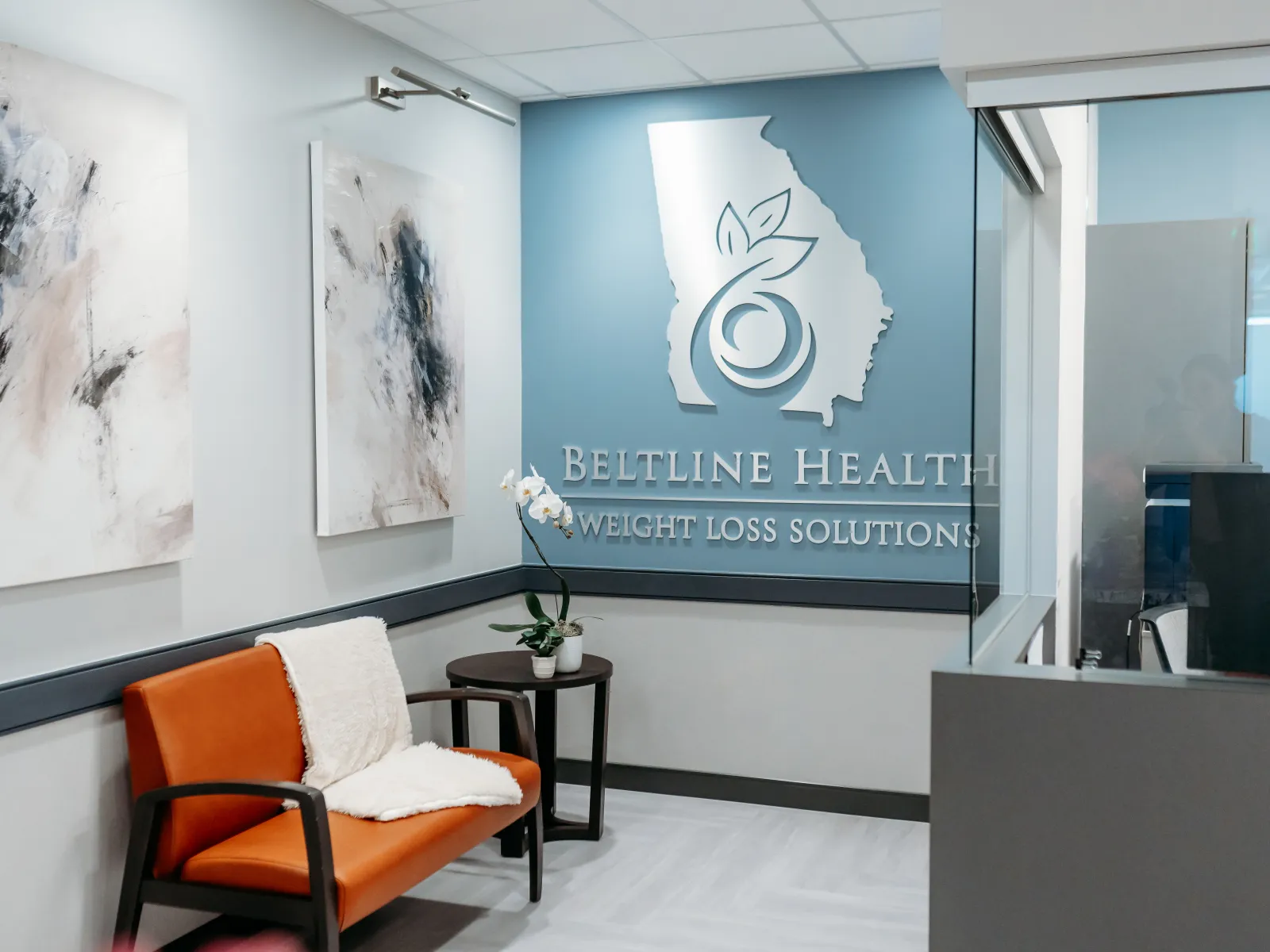 Modern waiting room with orange chair, abstract wall art, and Beltline Health Weight Loss Solutions logo on blue wall.