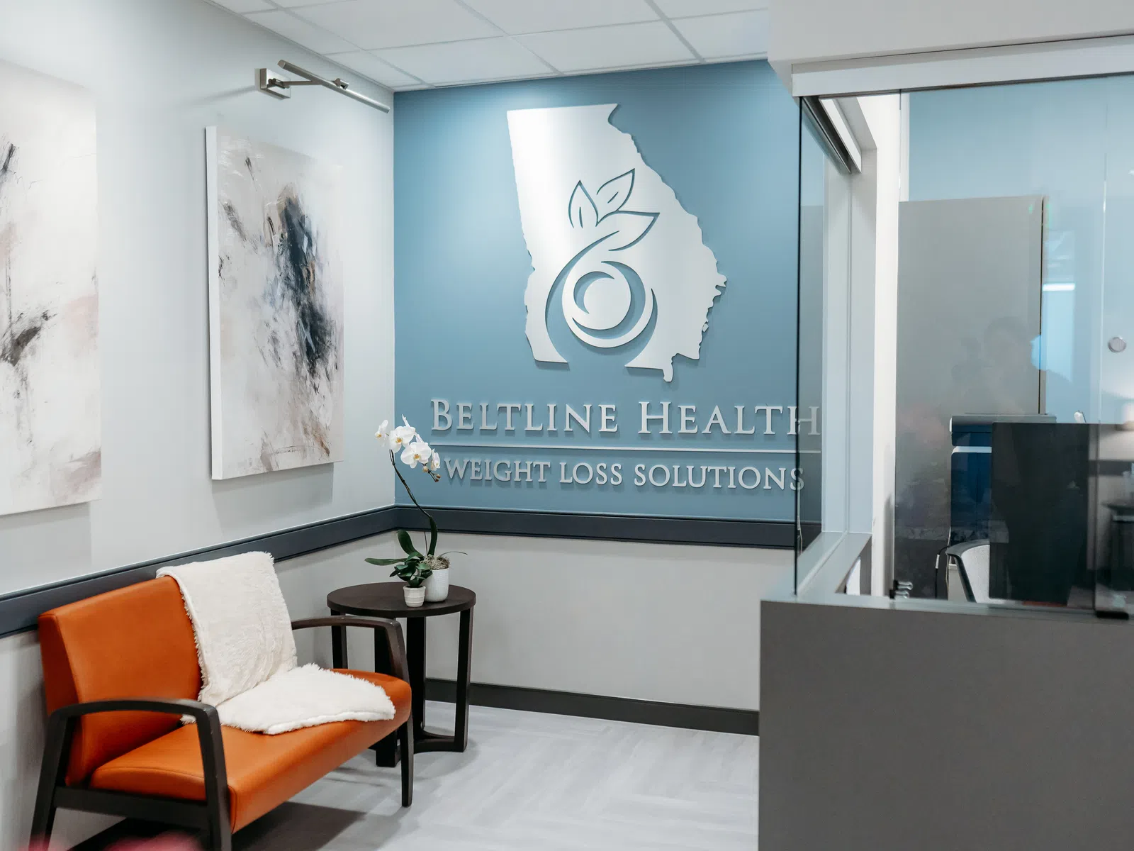 Modern waiting room with orange chair, abstract wall art, and Beltline Health Weight Loss Solutions logo on blue wall.