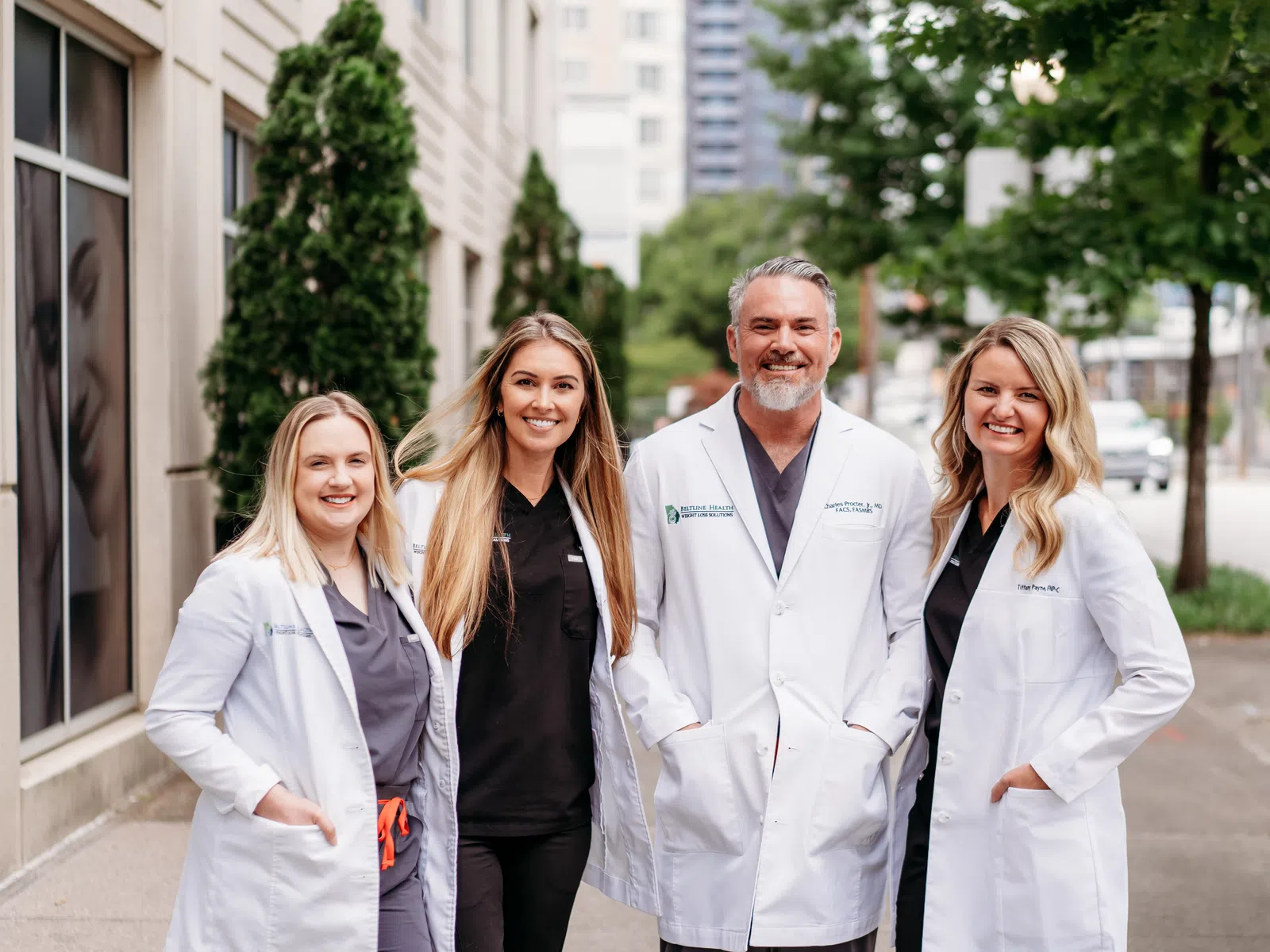 Group of four smiling healthcare professionals wearing white coats outdoors in an urban setting.