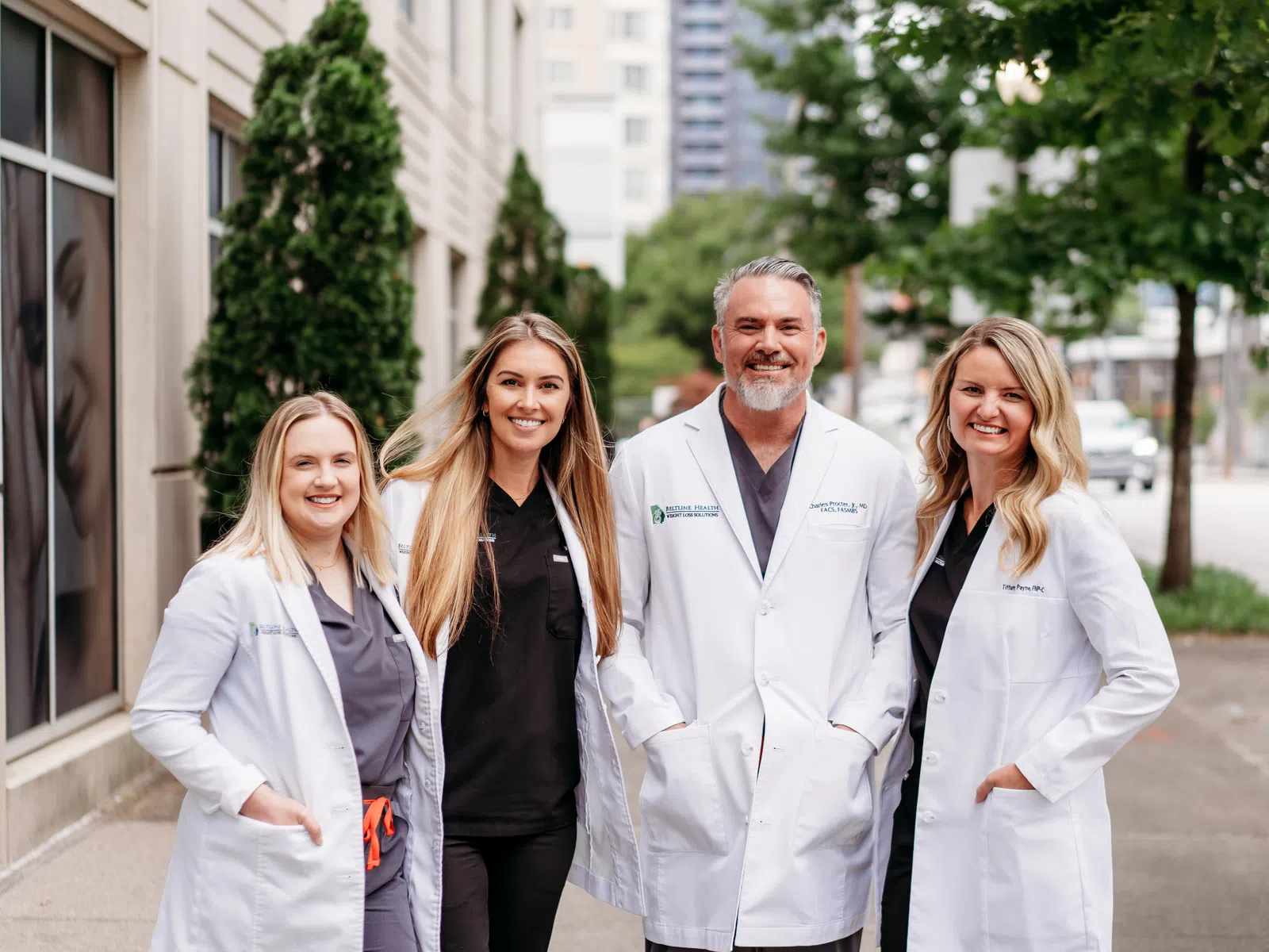 Group of four smiling healthcare professionals wearing white coats outdoors in an urban setting.