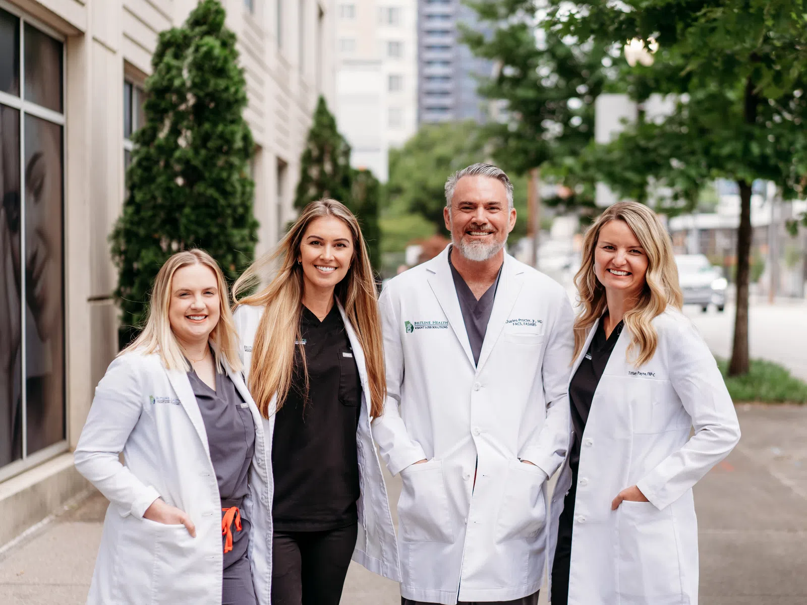 Group of four smiling healthcare professionals wearing white coats outdoors in an urban setting.