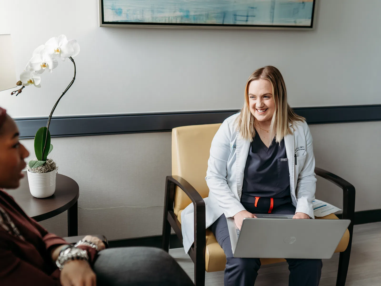 Female doctor with laptop smiling and consulting a patient in a modern medical office with orchid plant.