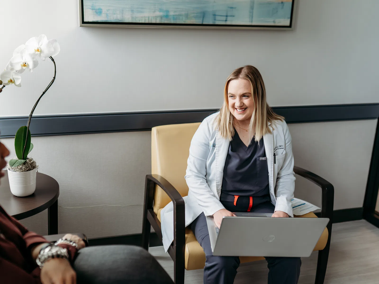 Female doctor with laptop smiling and consulting a patient in a modern medical office with orchid plant.