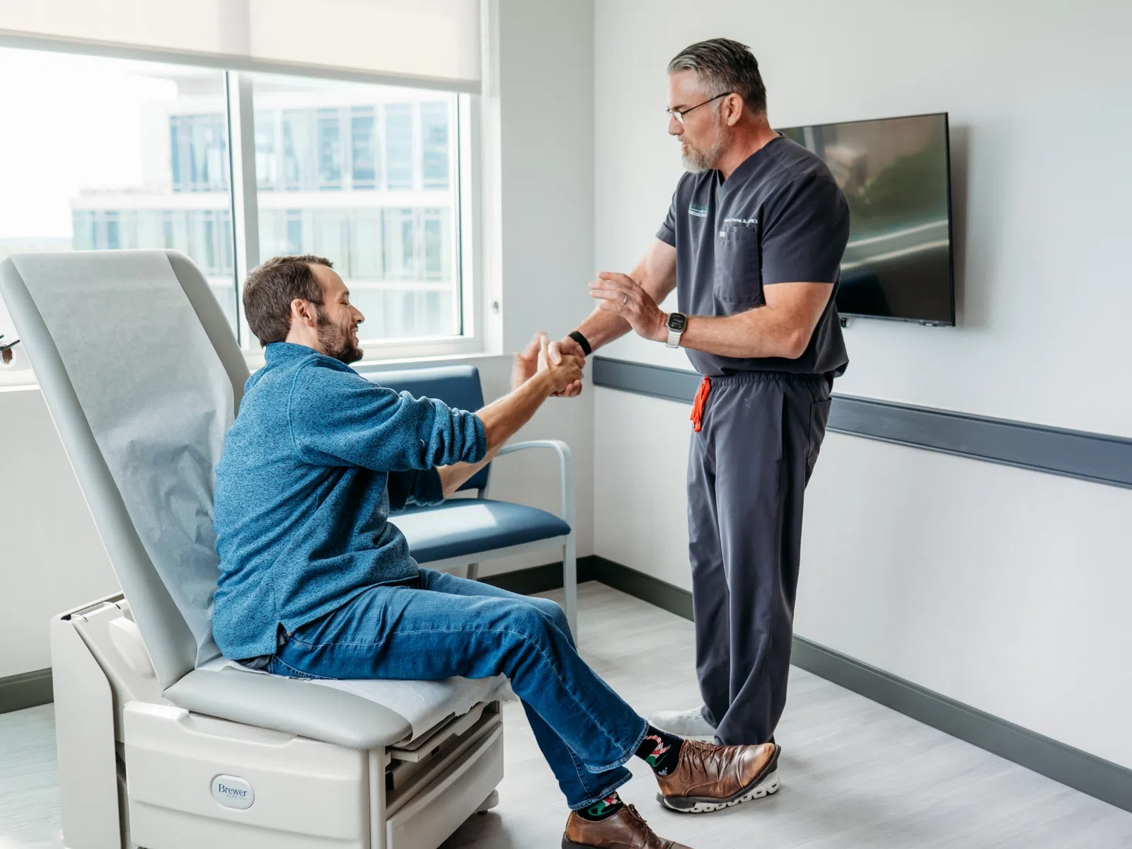 Physical therapist assisting man seated on exam table with wrist stretch in modern clinic room.