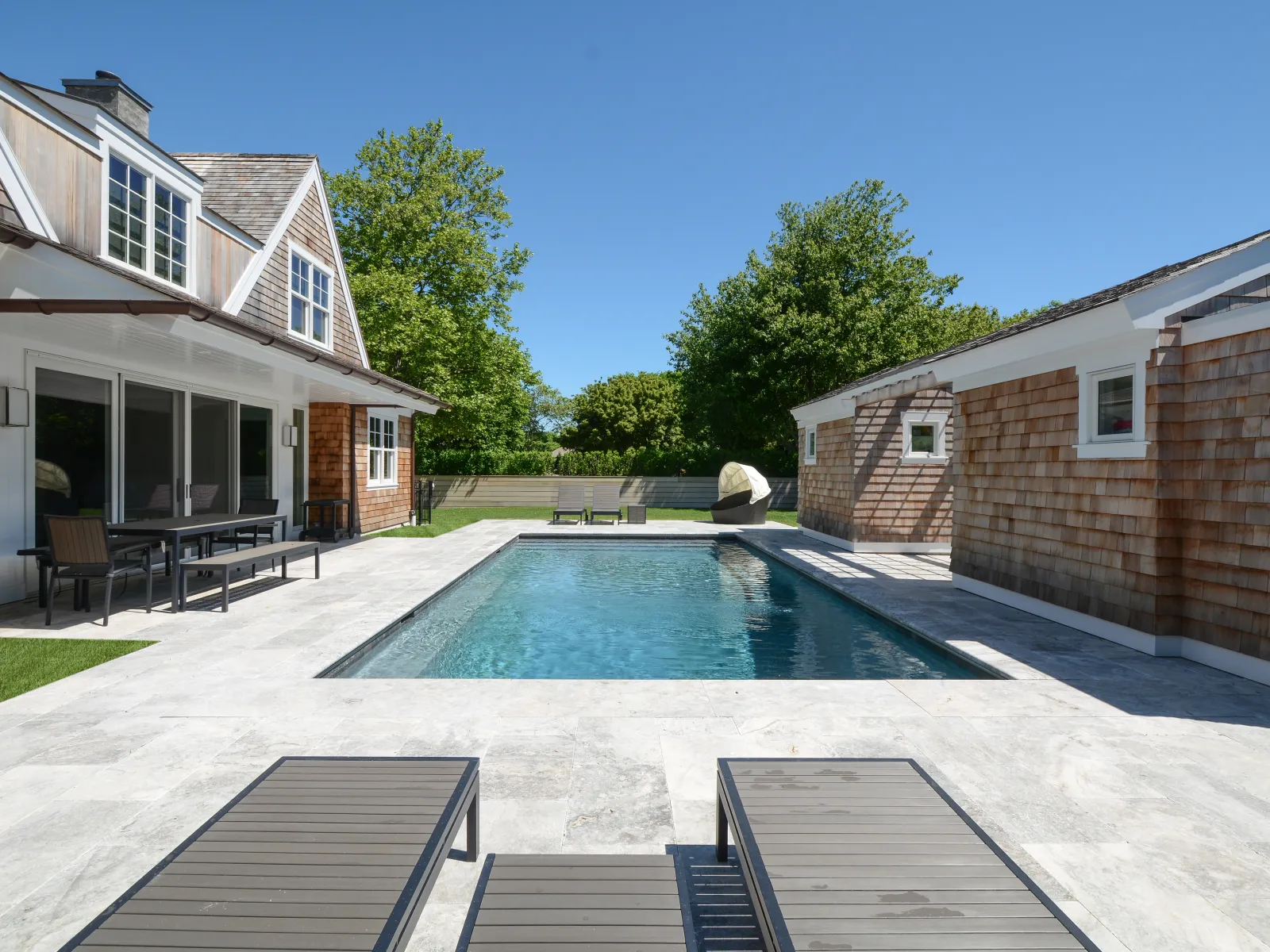 Modern backyard with a rectangular swimming pool, lounge chairs, outdoor dining area, and two shingle-style houses.
