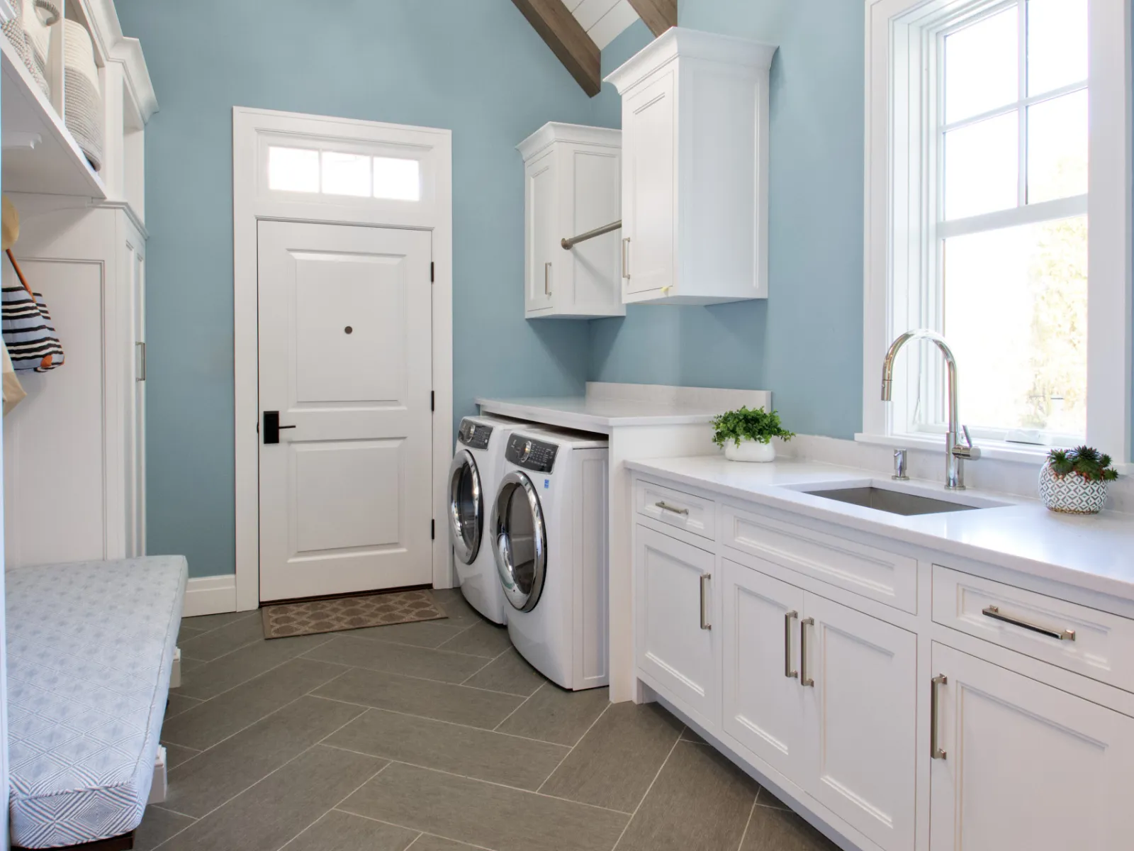 Bright laundry room with white cabinets, front-load washer and dryer, blue walls, vaulted ceiling, and modern light fixture.