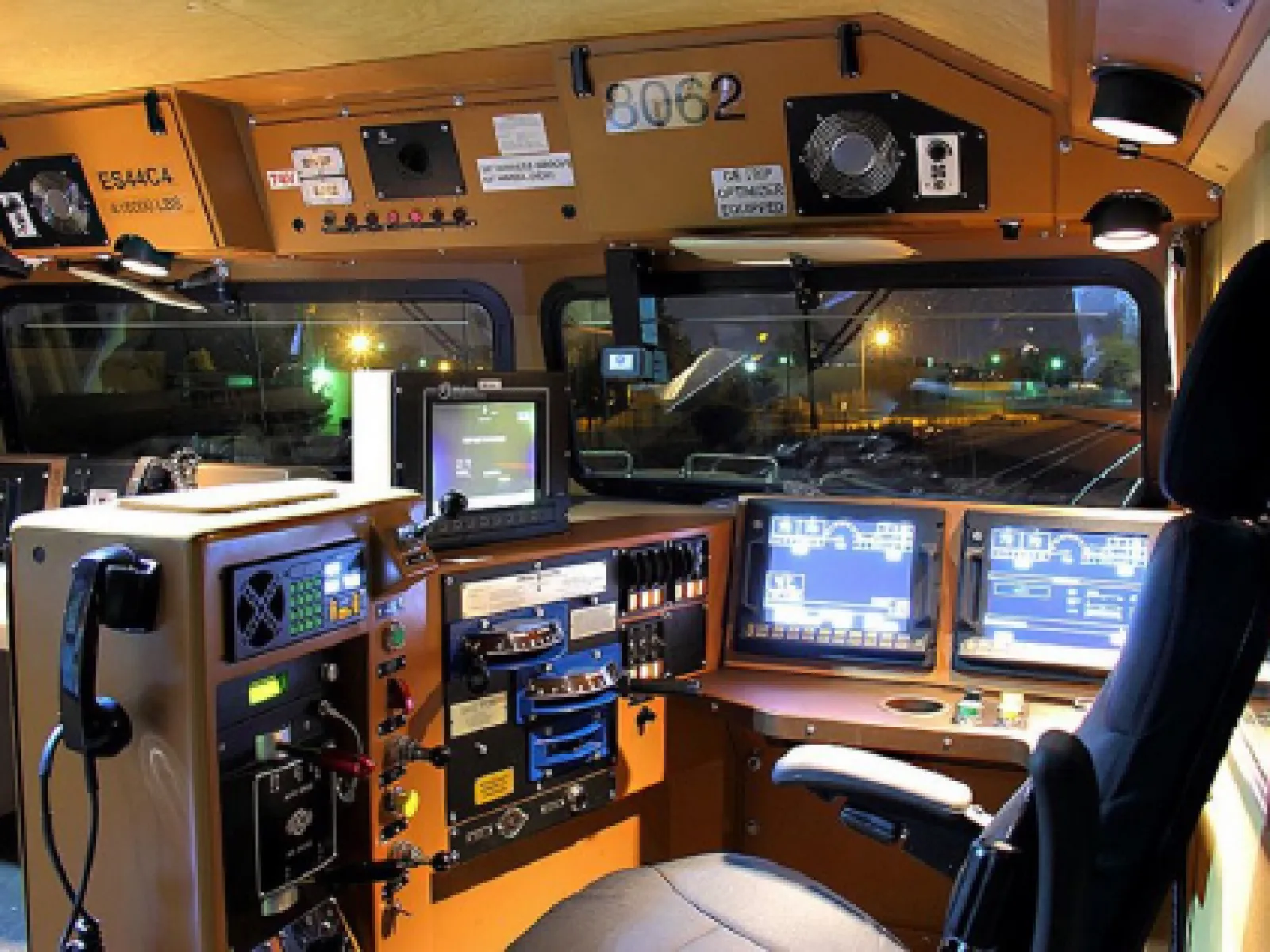 Train engineer cab interior with control panels, monitors, communication devices, and a driver seat at night.