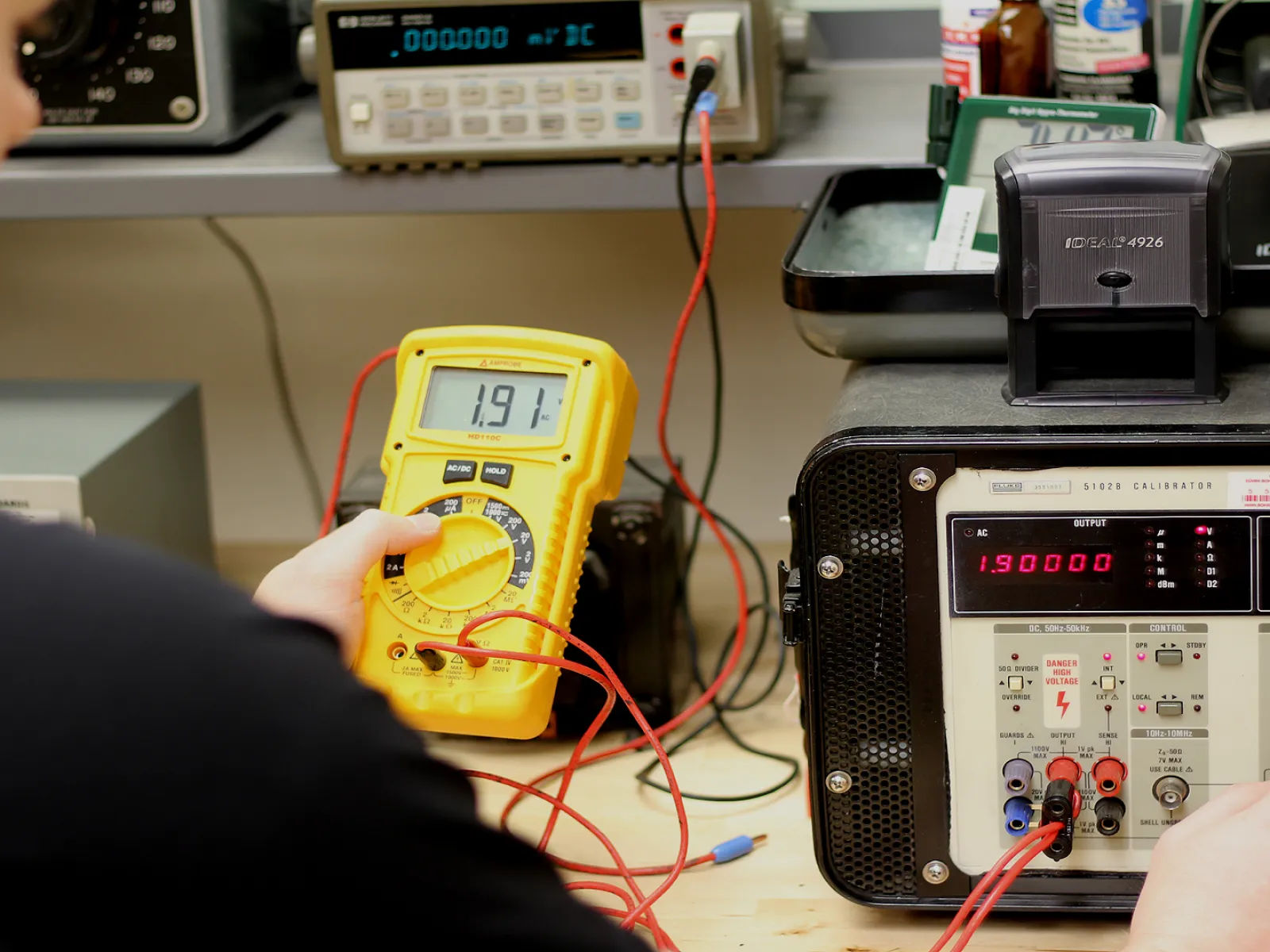 Person testing electrical equipment using a yellow multimeter and precision calibration instruments on a workbench