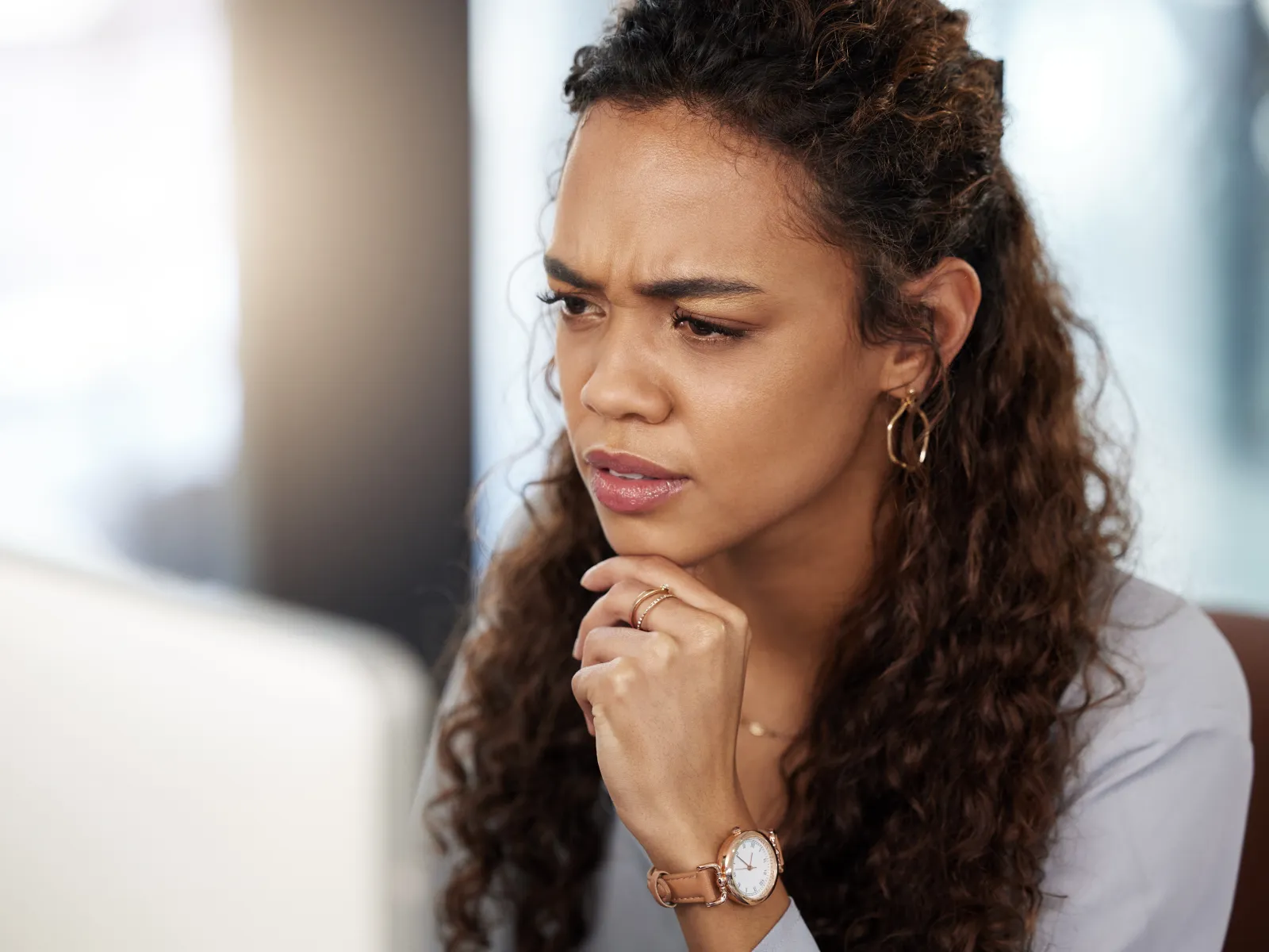 Young woman with curly hair looking confused while working on a laptop in a bright room.