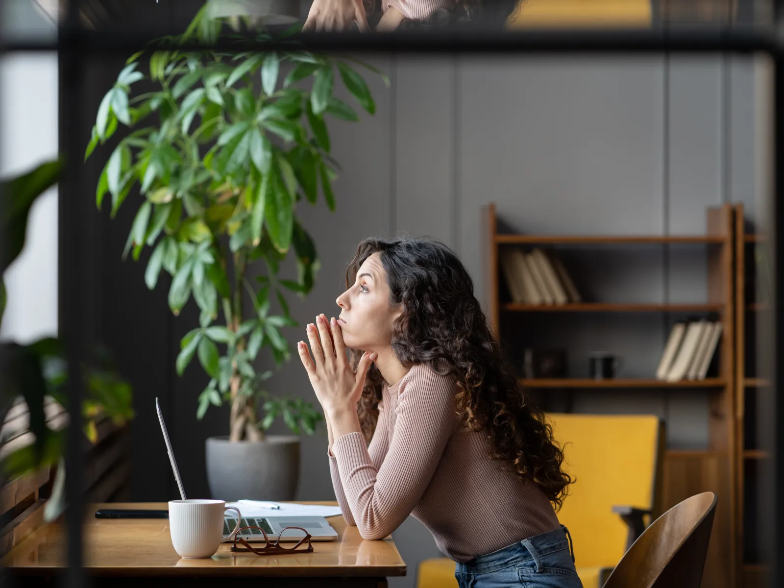 Woman sitting at desk in modern office, looking thoughtfully out the window with hands clasped near face.