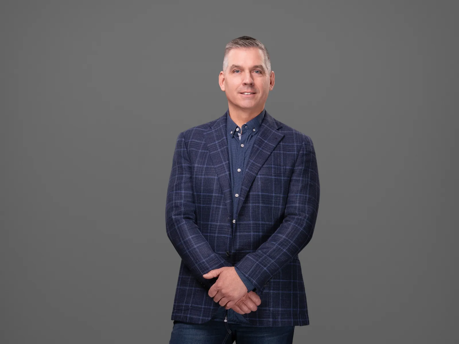 Man in a blue checked blazer and jeans standing confidently against a gray background