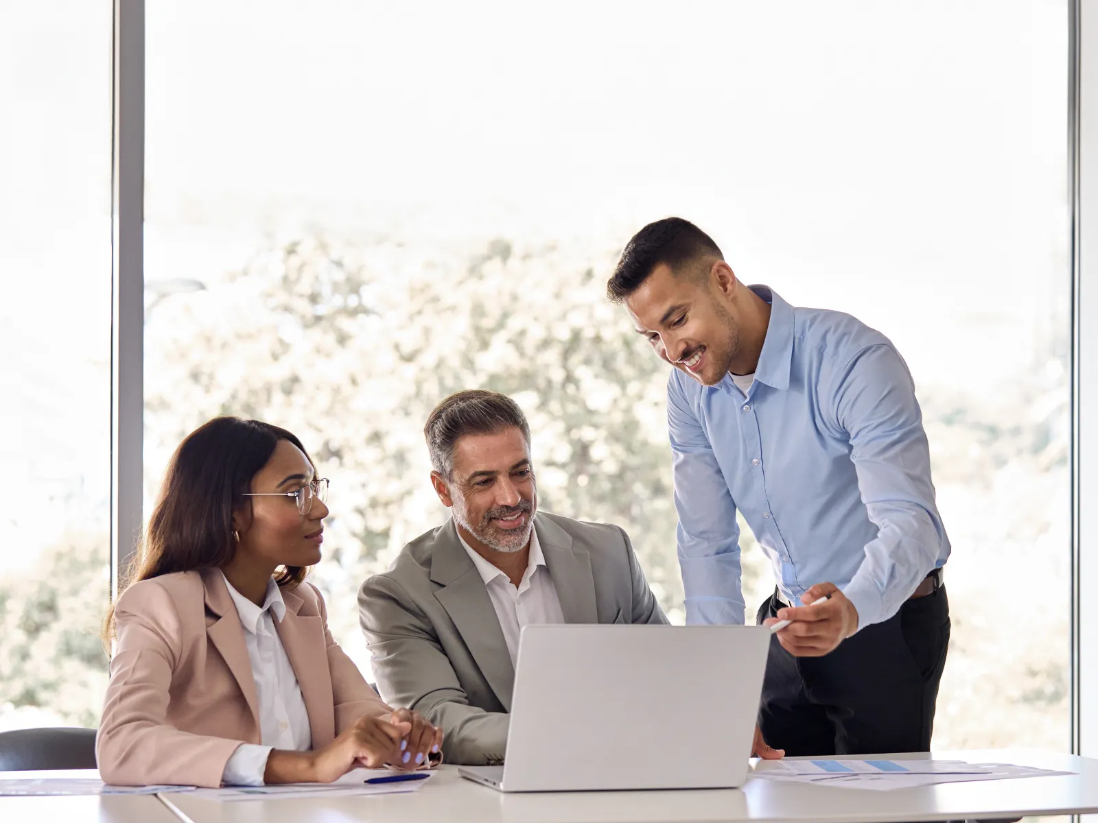 Three diverse professionals collaborating over a laptop in a bright modern office with large windows.