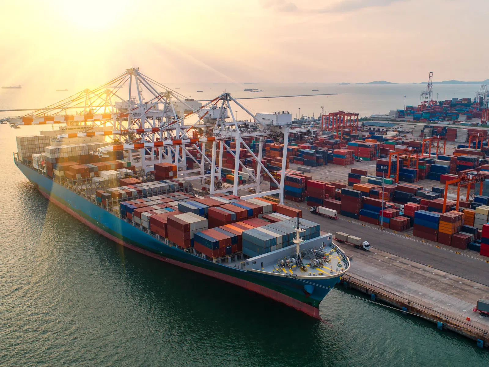 Cargo ship loaded with containers docked at a busy port with cranes during sunset over calm water.