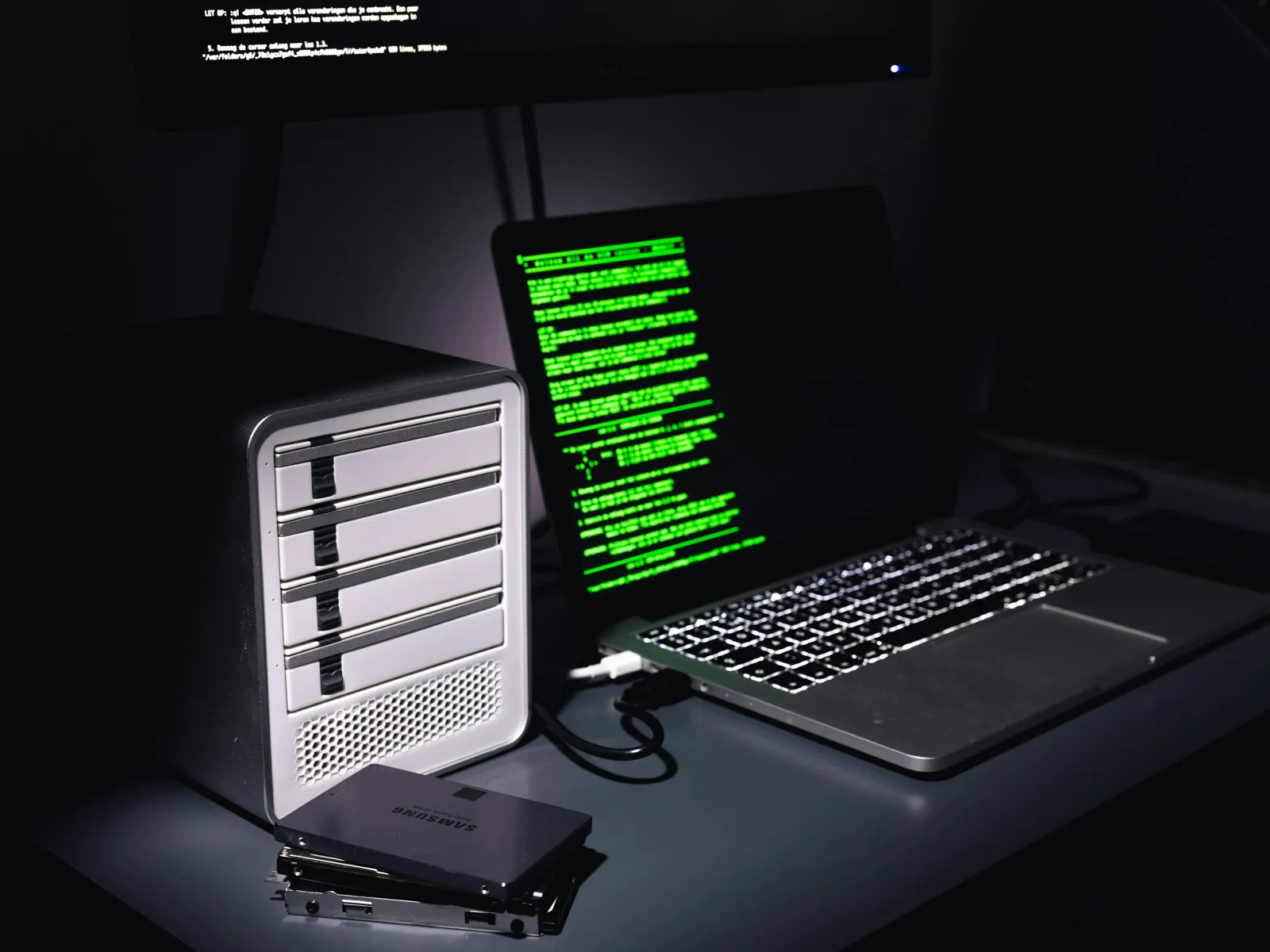 Laptop with green code on screen connected to external storage drives on dark desk setup.