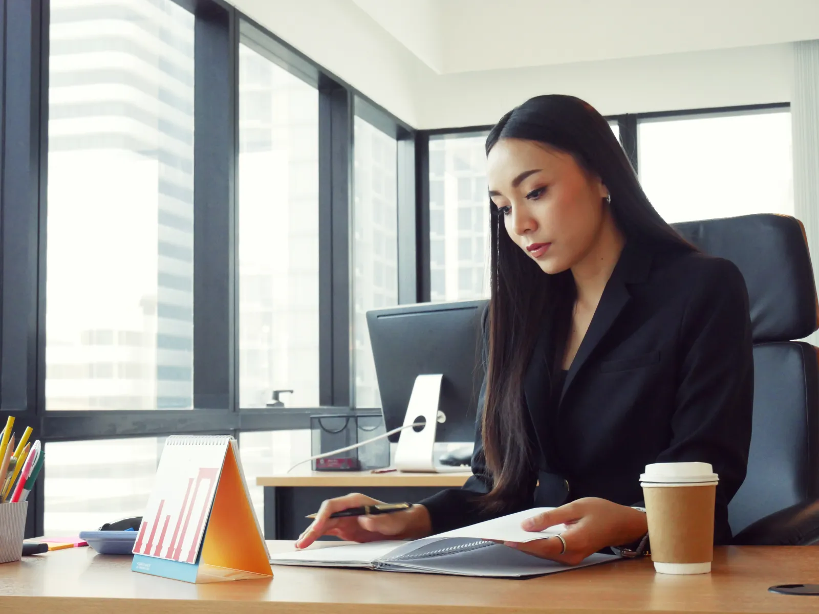 Businesswoman reviewing documents at office desk with coffee and charts near large windows in daytime