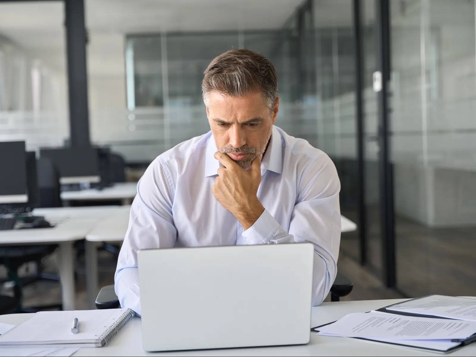 Professional man in a white shirt concentrating on laptop work in a modern office with documents and notebook.