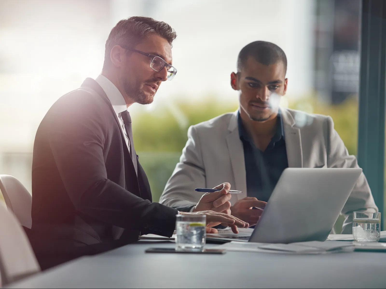 Two professional men in suits discuss work at a laptop in a bright office meeting room with glasses of water.
