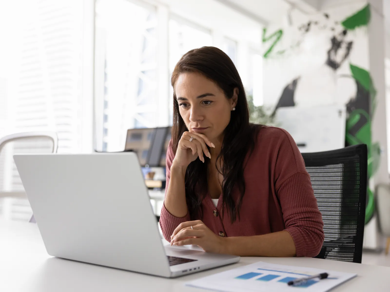 Woman focused on laptop screen while working in a bright modern office with charts on the desk.