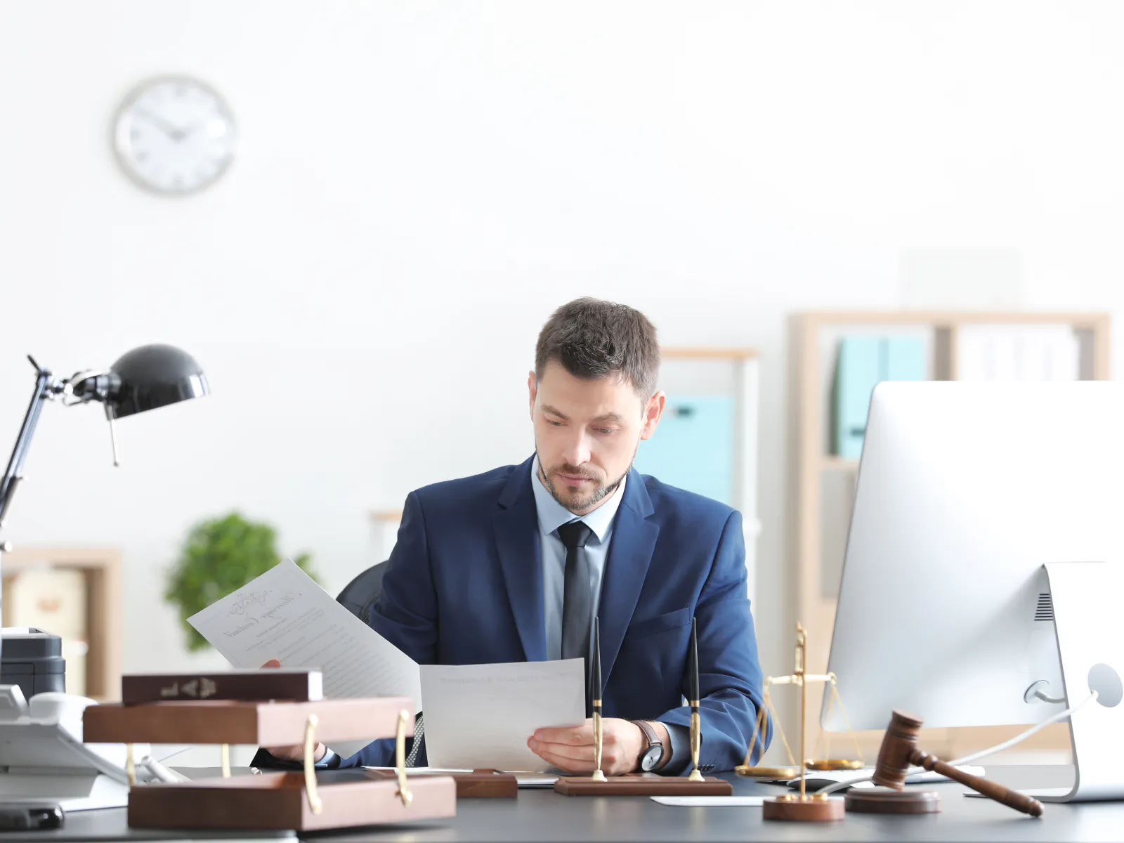 Professional man in suit reviewing documents at office desk with computer, lamp, and legal accessories.