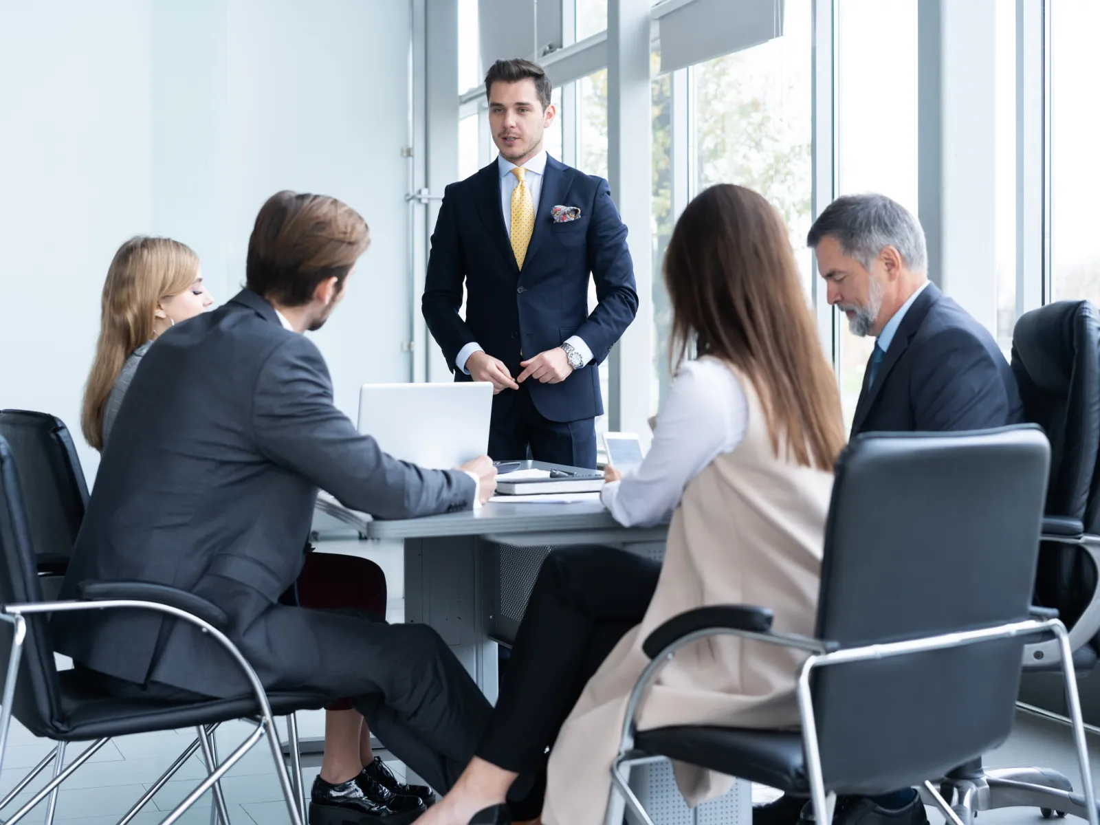 Businessman presenting in a modern meeting room to colleagues seated around a conference table with laptops.