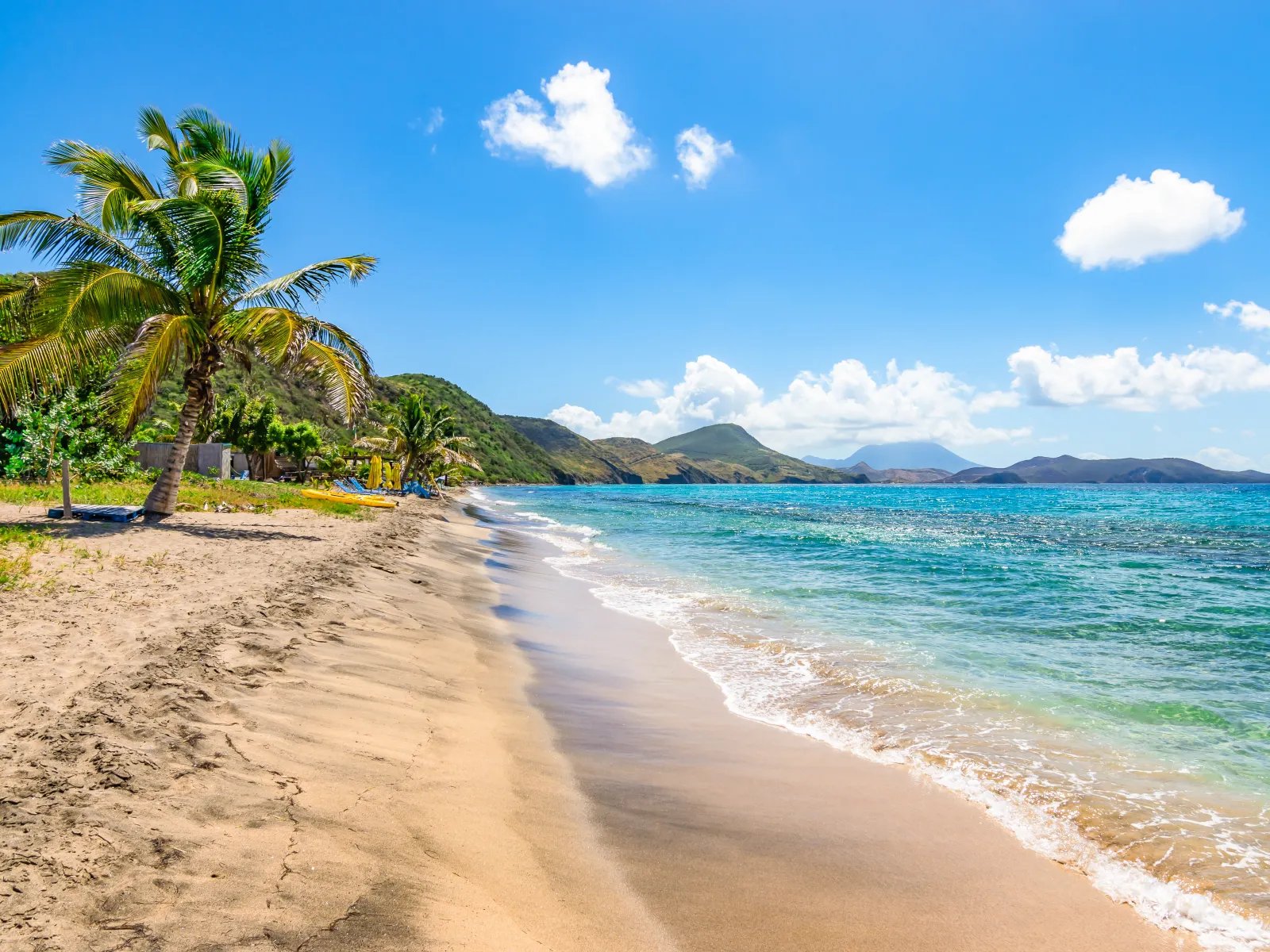 Sunny tropical beach with palm trees, golden sand, clear blue sea, and distant hills under a bright sky.