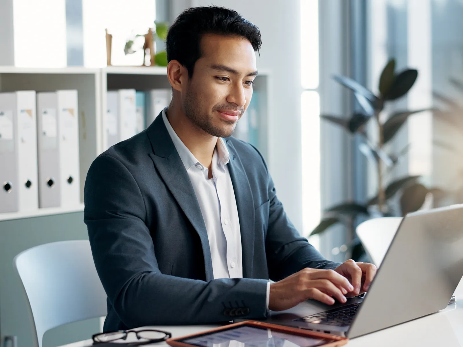 Young businessman in blazer working on laptop at modern bright office desk with tablet and glasses.