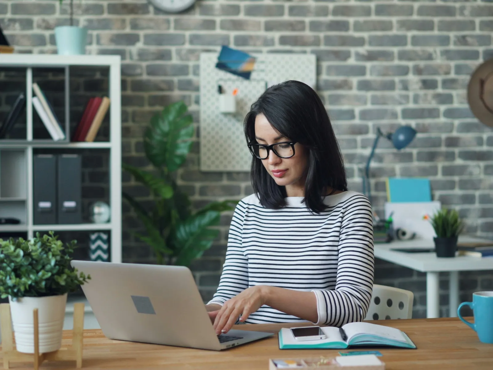 Woman in glasses working on laptop at wooden desk with plants and open book in modern office space.