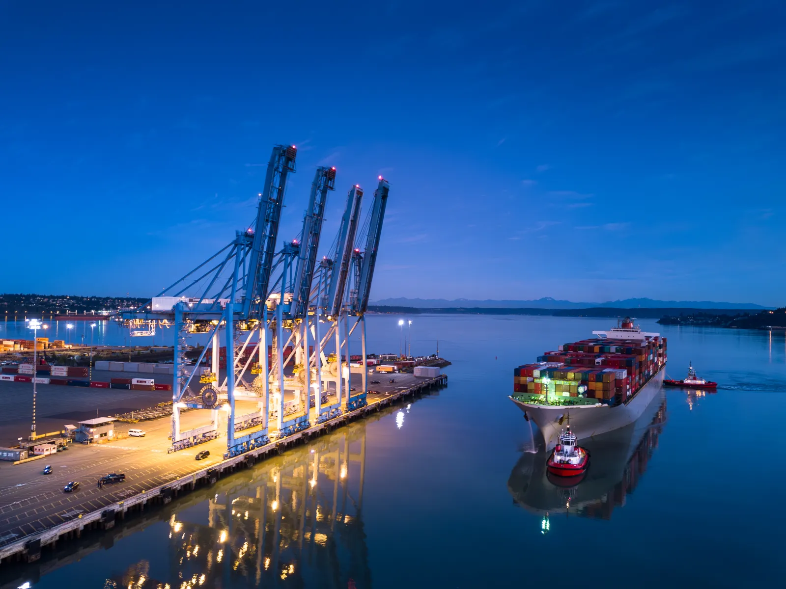 Container ship docked at illuminated port cranes during twilight with calm water reflections and tugboats nearby