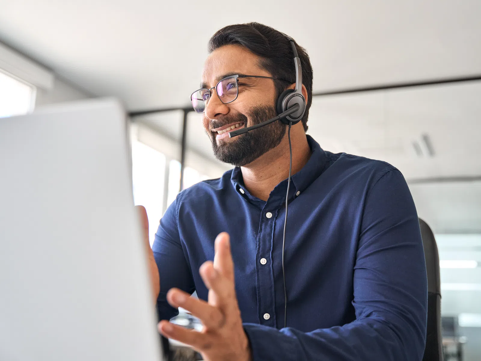 Smiling man in glasses and headset engaged in video call at modern office workspace