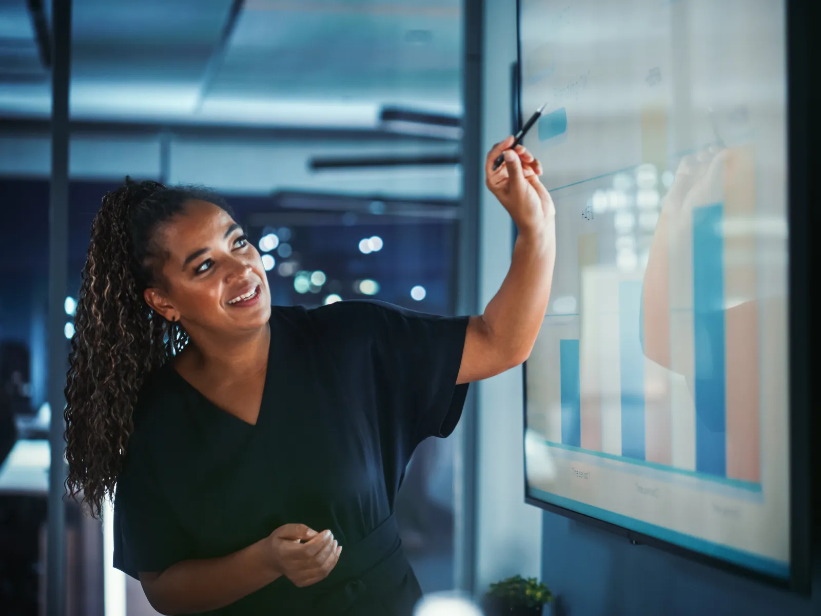 Smiling businesswoman presenting growing bar chart on digital screen in modern office at night