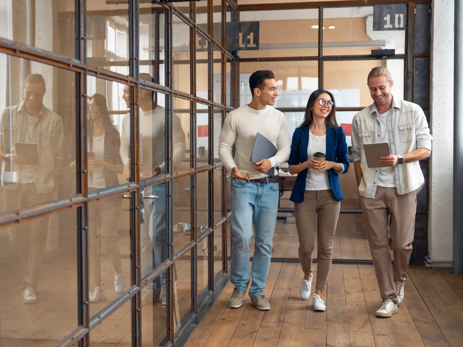 Three young professionals walking in an office hallway with laptops and coffee, smiling and chatting