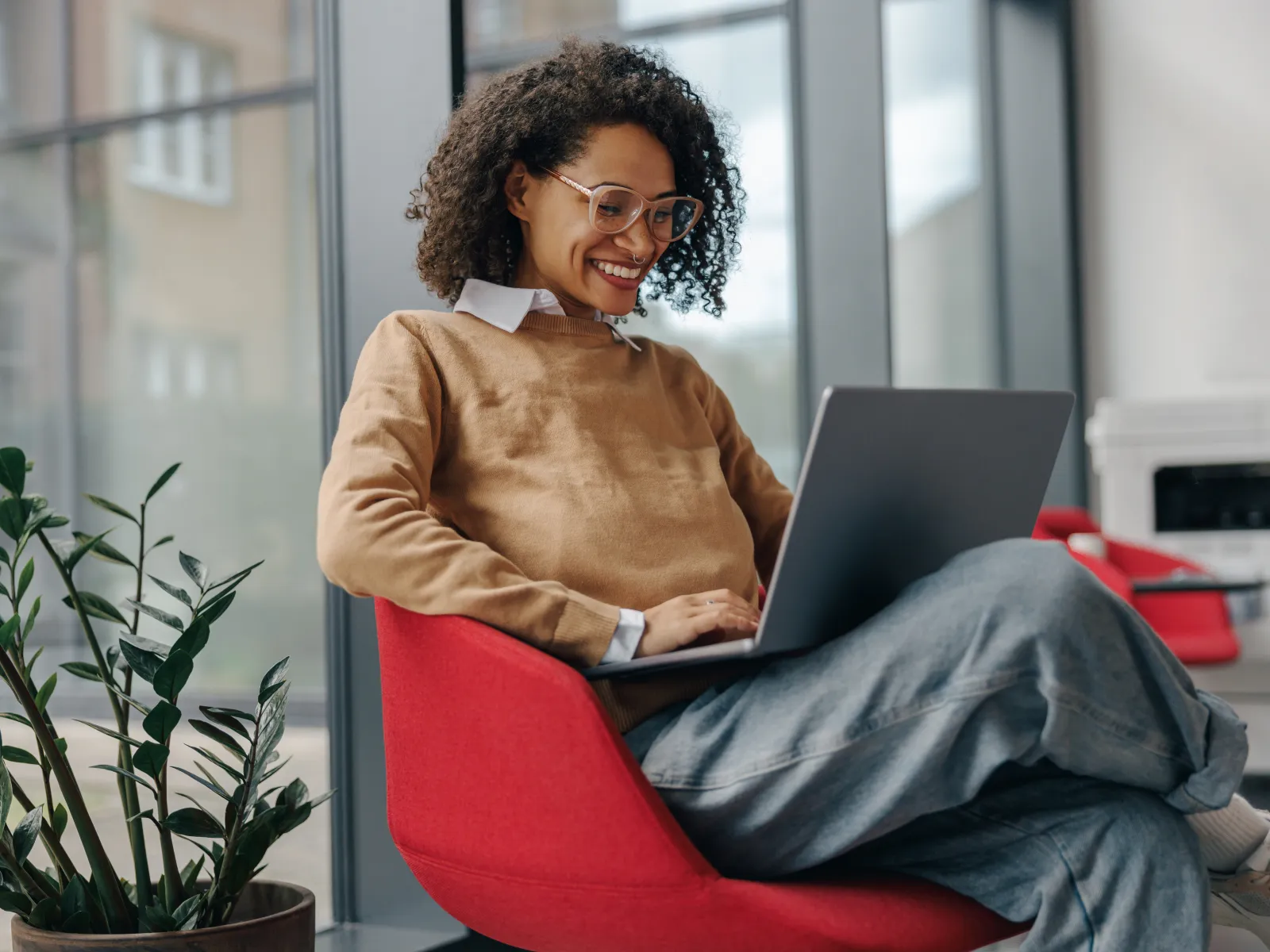 Smiling woman with curly hair and glasses working on laptop seated in red chair beside indoor plant