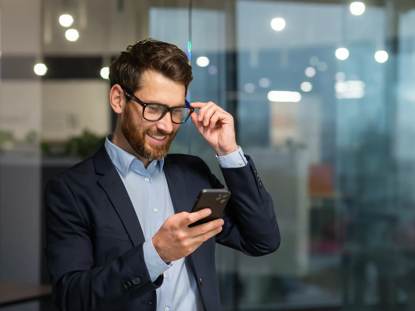 Smiling man in business attire adjusts glasses while using smartphone in modern office setting