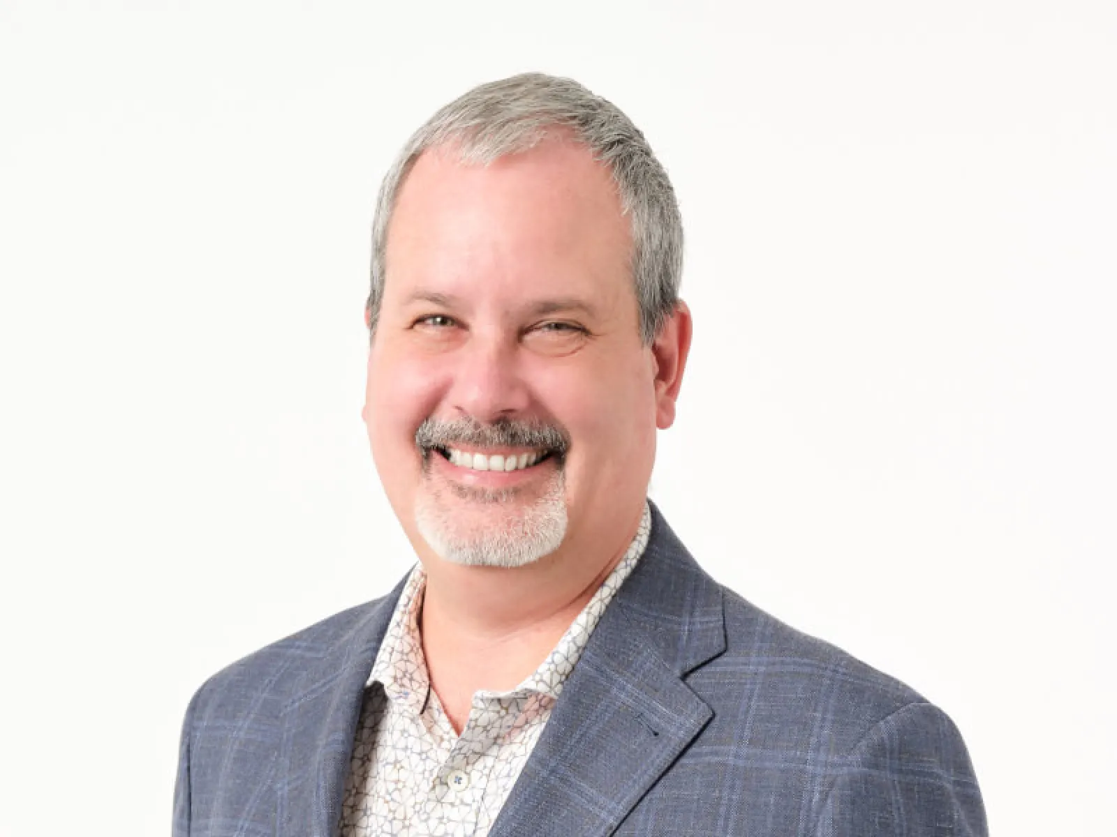 Smiling middle-aged man with gray hair and a goatee wearing a checkered blazer and patterned shirt on white background