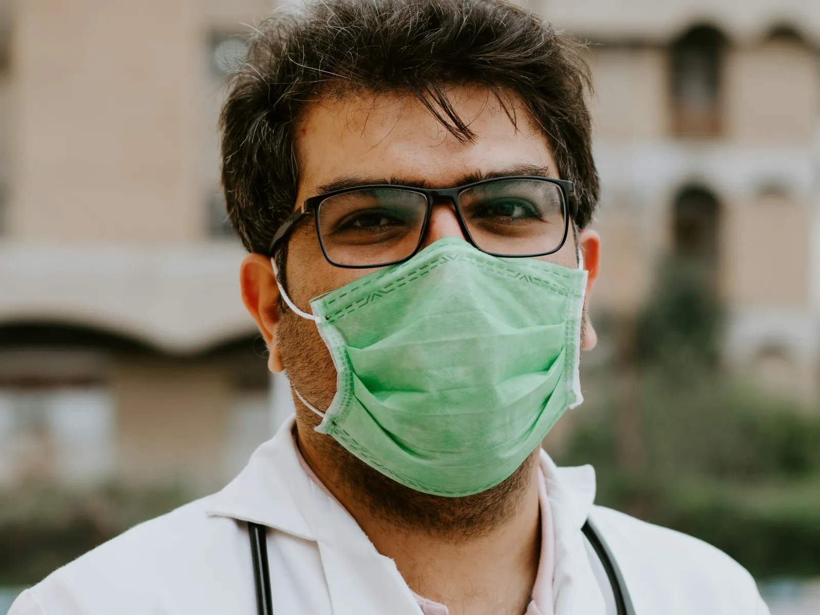 Male doctor wearing a green face mask, glasses, white coat, and stethoscope standing outdoors near a building.