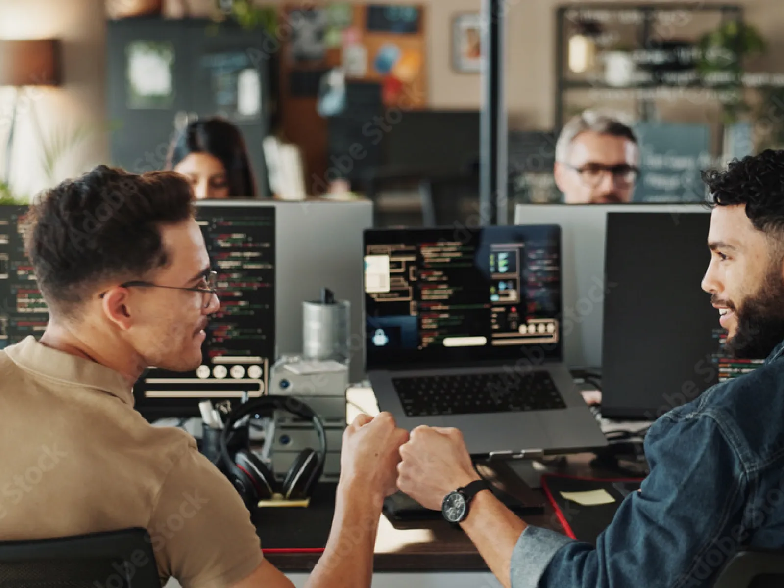 Two men fist bumping at a tech workspace with multiple screens displaying code and data.