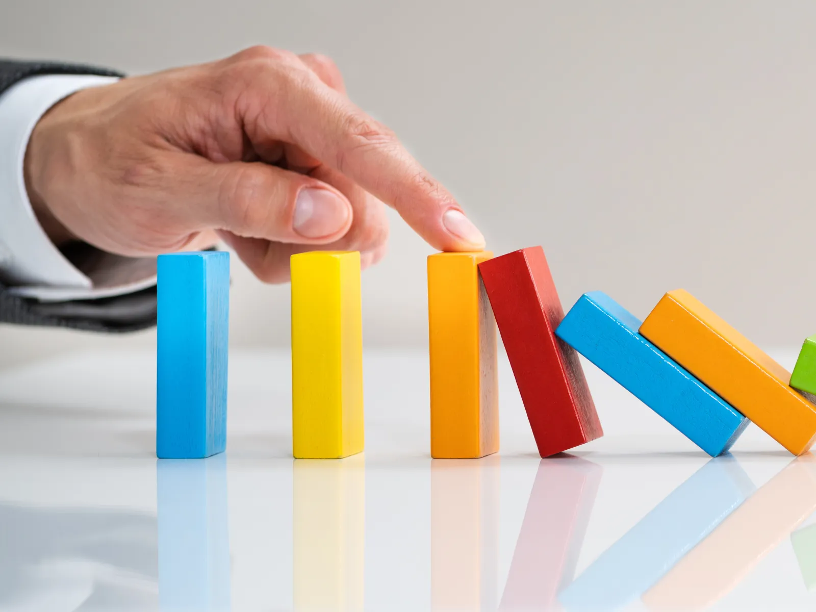 Hand stopping colorful dominoes from falling on a white reflective surface representing control and prevention.