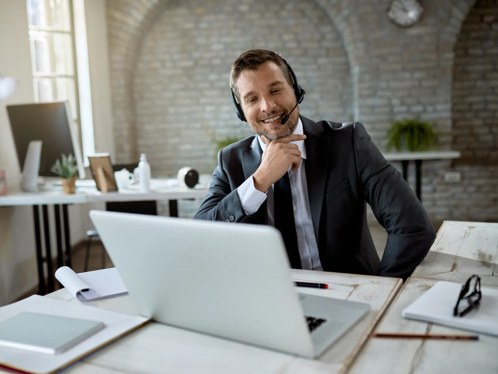Smiling businessman in headset having video call with laptop in bright modern office with brick walls.