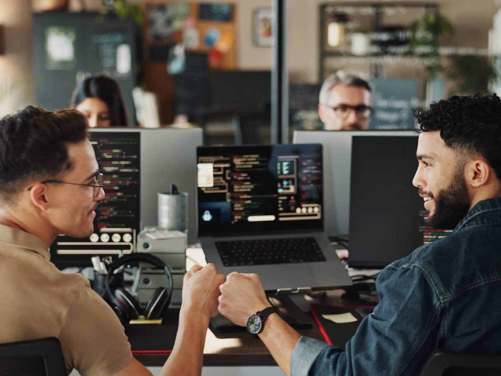 Two software developers fist bump while working on coding projects at a shared office workspace.