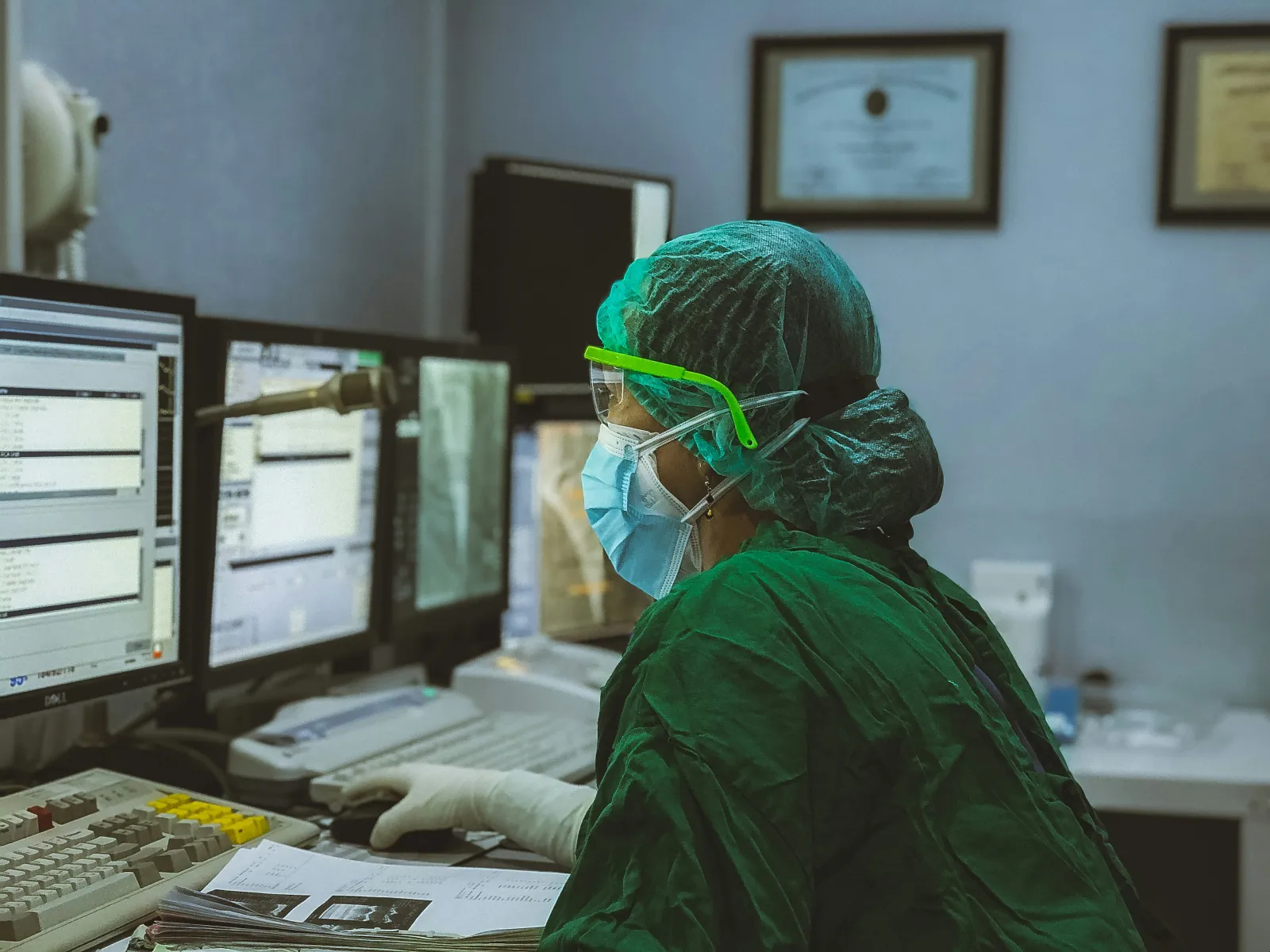 Medical professional in protective gear analyzing data on multiple computer screens in a clinical setting.