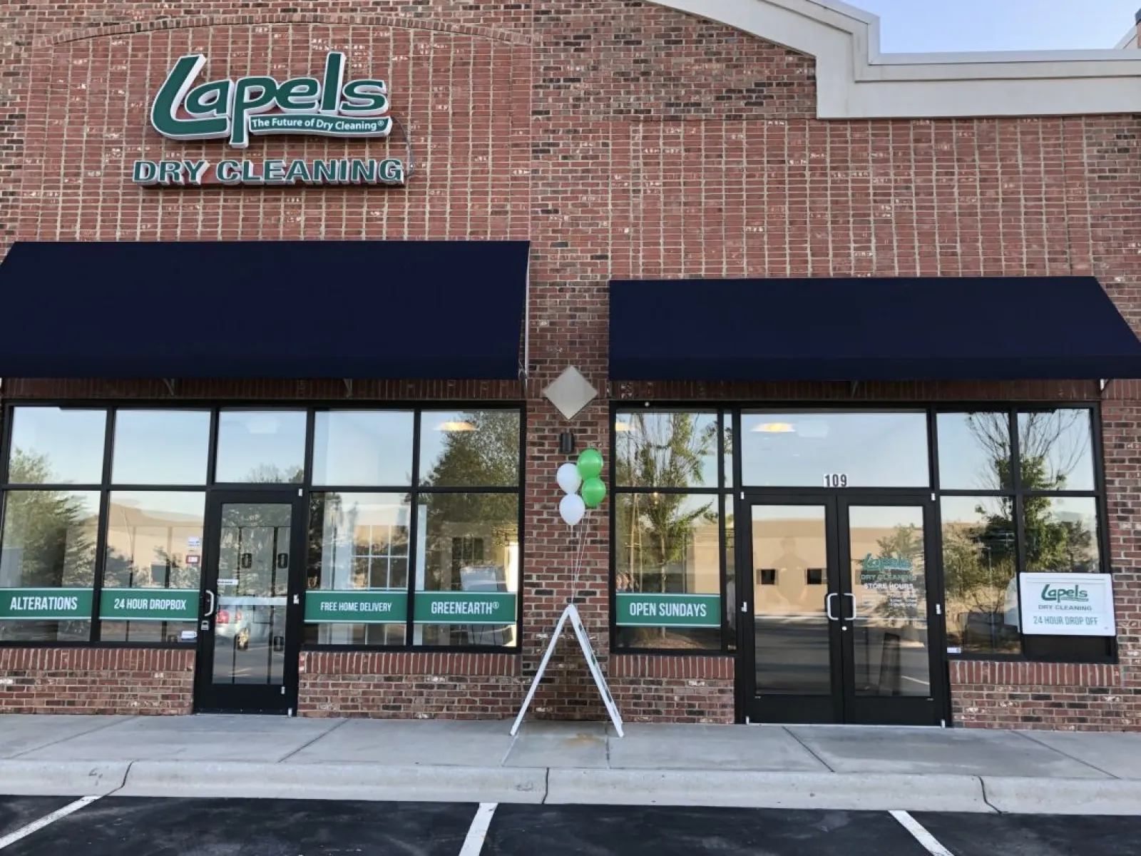 Brick facade of Lapels Dry Cleaning store with black awnings, balloons, and signs advertising services and Sunday hours