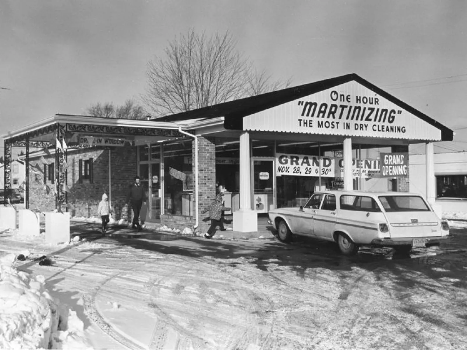 Black and white photo of a dry cleaning business with a car parked outside and people entering during snowy winter.