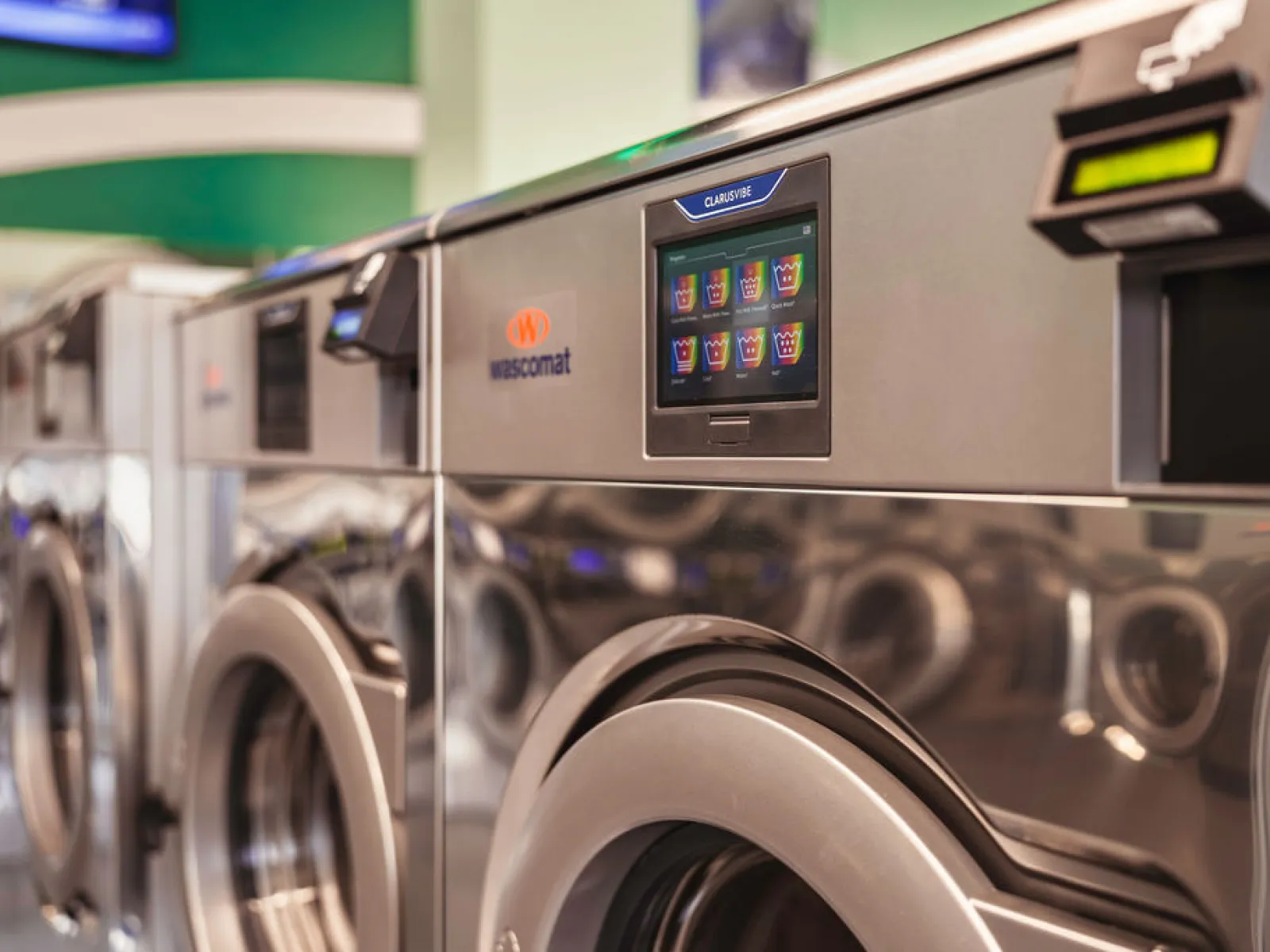 Row of modern Wascomat commercial washing machines with digital touchscreens in a laundromat.