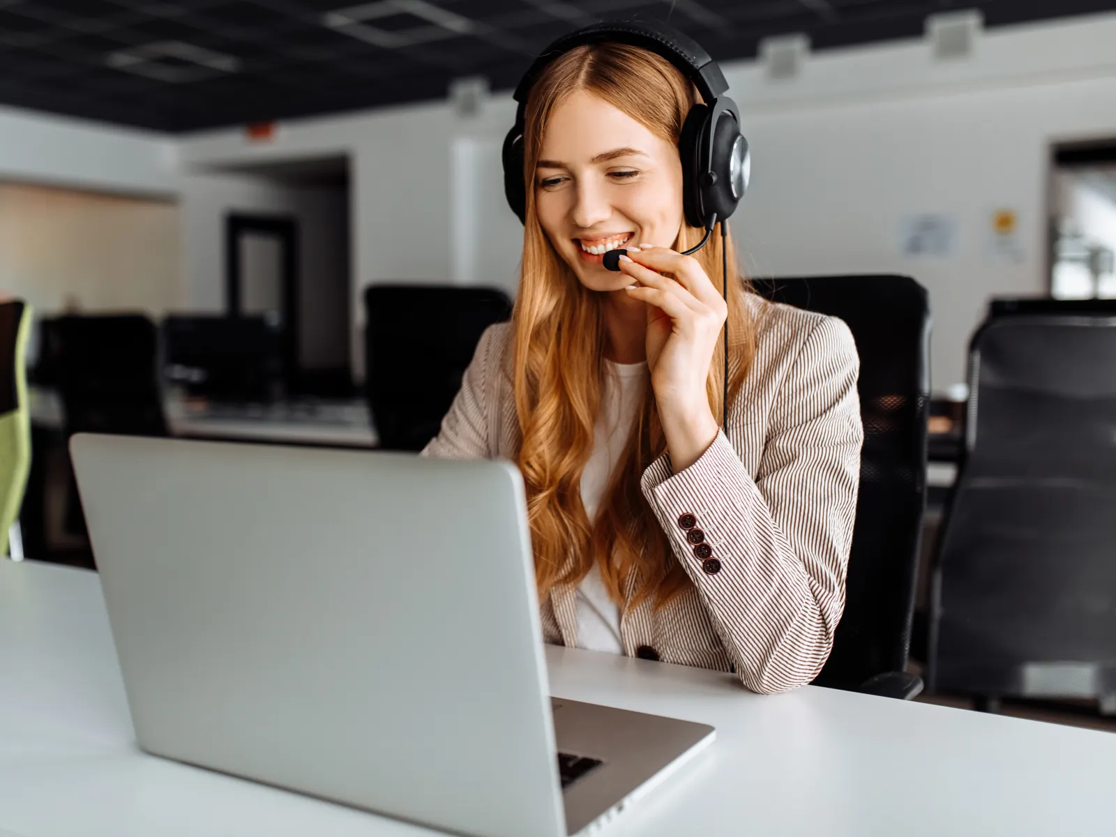 Young woman with headset smiling and working on laptop in modern office environment