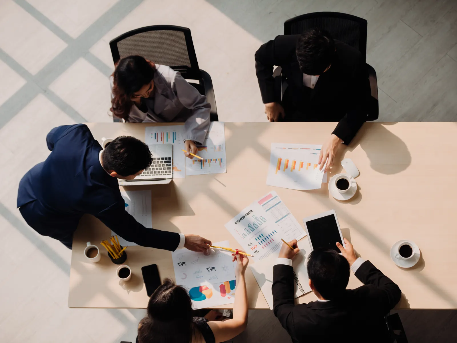 Top view of business team discussing charts and data on a desk with laptops, tablets, and coffee cups