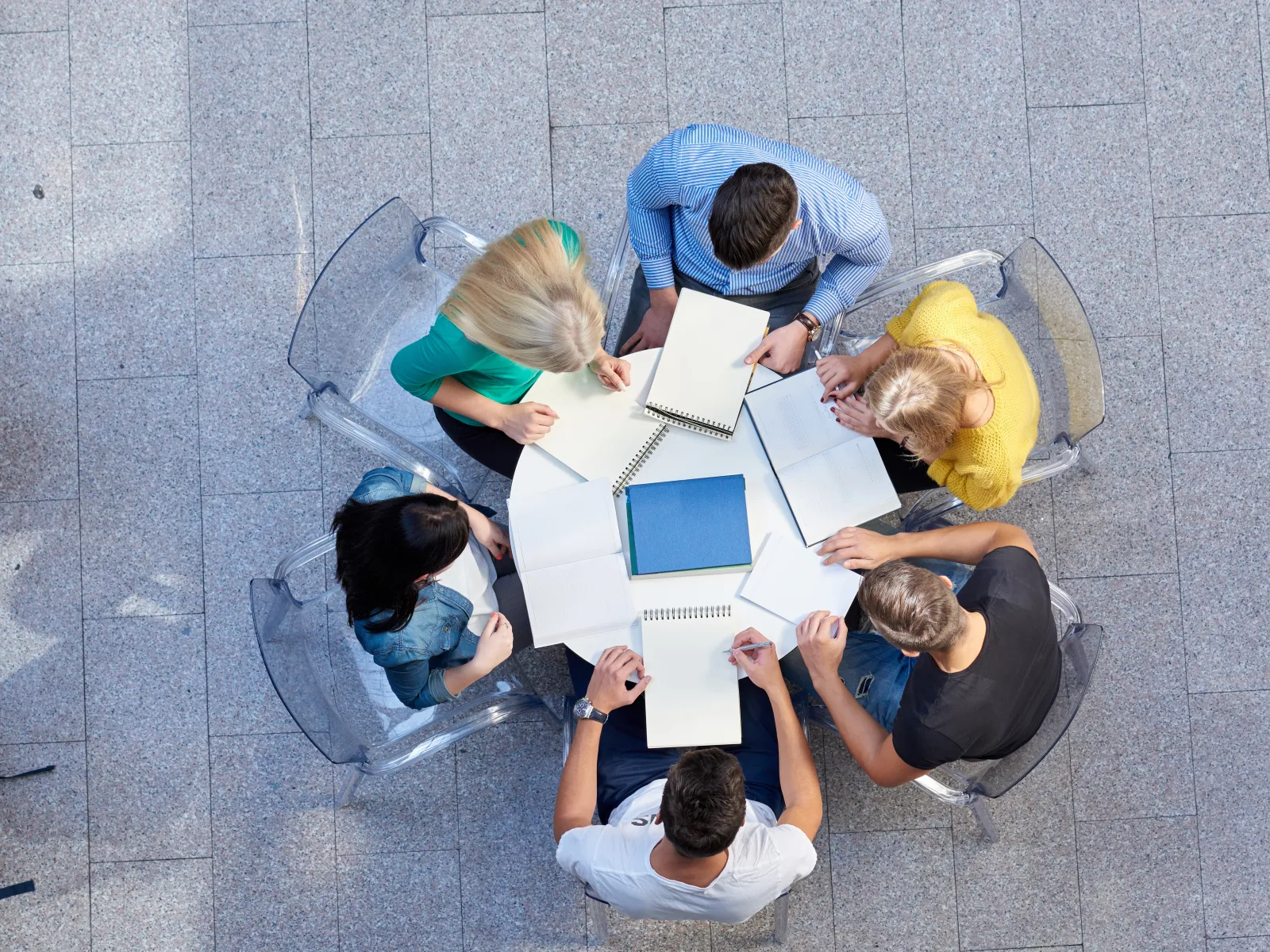 Overhead view of a diverse group studying together with notebooks around a round white table on tiled floor.